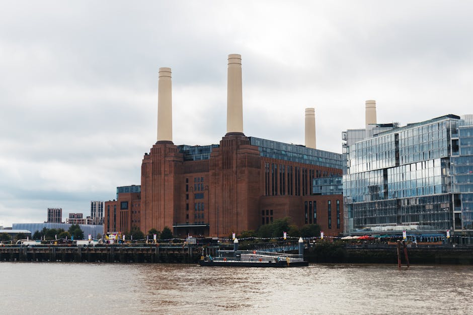 The image depicts a large industrial building with a historic red brick facade, featuring tall, cream-colored smokestacks extending upward. Adjacent to the building, there is modern glass-fronted office or commercial space with reflective windows, creating a contrast between old and new architecture. The scene is situated along a waterfront, with calm brownish water in the foreground and a small boat docked close to the shore. The sky above is overcast with grey clouds, contributing to a subdued lighting that illuminates the scene evenly. In the surrounding area, some greenery including trees and bushes can be seen, but the focus remains on the industrial building and modern structure, emphasizing the urban landscape. The image captures a mix of historical industrial and contemporary architectural elements, set against a waterway with minimal human activity visible.