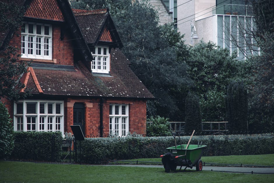 A residential garden scene featuring a well-maintained lawn with lush green grass, bordered by neatly trimmed hedges and a variety of leafy trees providing shade. In the foreground, a green wheelbarrow parked on a paved pathway contains gardening tools such as a rake and shovel, indicating active garden maintenance. To the side, a black garden chair is placed near the hedge, suggesting outdoor relaxation. The background showcases a traditional red-brick house with white-framed, multi-pane windows, a tiled roof with visible moss patches, and small dormer windows. Tall modern buildings can be seen behind the trees, illustrating an urban garden setting. The garden is damp, with slightly wet soil and tender leaves, suggesting recent watering or rain. Overall, the scene depicts a tidy, well-kept outdoor space in the process of or ready for ongoing garden work, aligned with landscaping and lawn care themes that might be discussed in a 'SW18 gardeners real cost and maintenance guide,' provided by Gardeners Wandsworth.